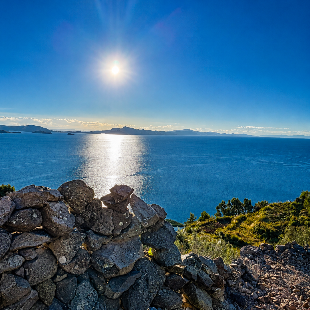 A scenic coastal landscape in Lake Titicaca with a bright sun over a deep blue sea, distant mountains on the horizon, and a foreground of stacked dark rocks and green shrubs.