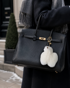 A black pebbled leather handbag with gold-tone hardware and a fluffy white pom-pom fur charm, carried by a person wearing a dark coat and gray scarf in an outdoor urban setting.