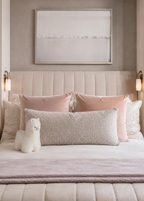 A beautifully styled bedroom featuring a bed with a textured beige pillow, blush pink pillows, a light mauve throw blanket, and a beautiful  white, fluffy alpaca plush made of alpaca fur. The background includes a gray wall with a framed print, built-in shelves, and a window overlooking a city skyline.