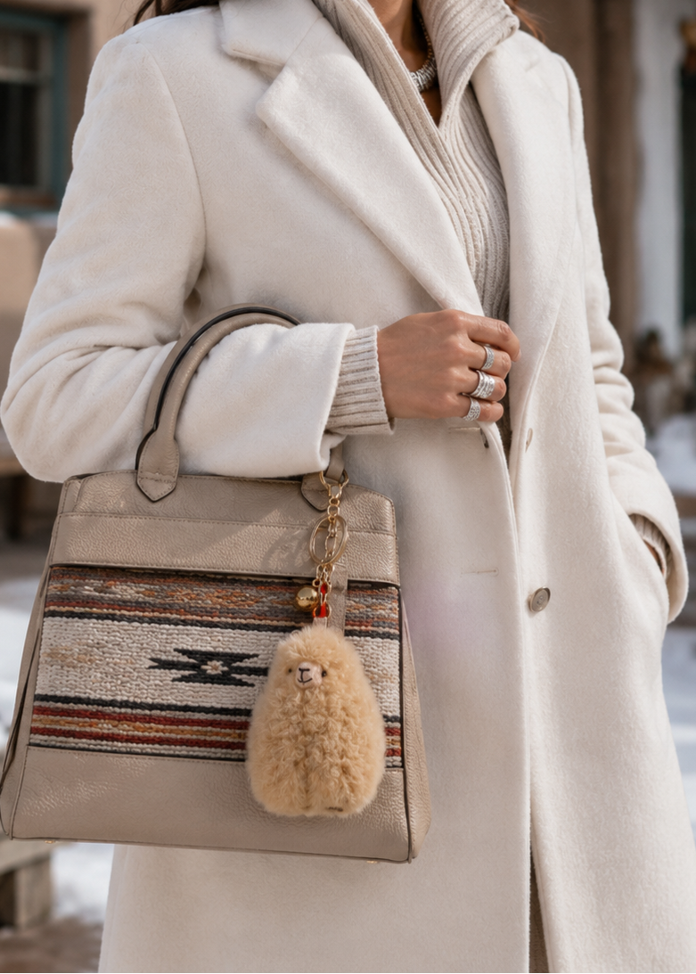 A woman wearing a long cream coat and ribbed cream sweater carries a beige woven handbag with a fluffy beige keychain charm in an outdoor snowy setting.