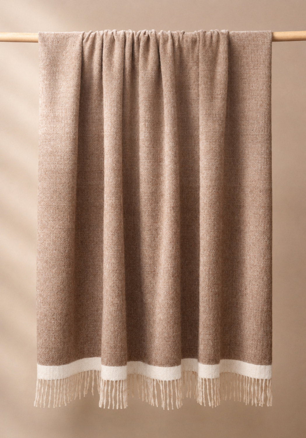 A draped woolen alpaca knitted throw blanket in a warm taupe-brown color, featuring a contrasting cream-colored band and fringe along the bottom edge, hung from a light wooden rod against a solid beige background.
