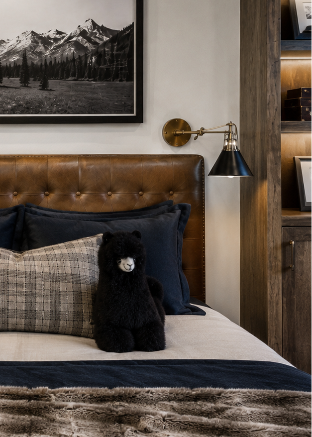A styled bedroom scene featuring a bed with a tufted brown leather headboard, dark navy pillows, a plaid accent pillow, a black plush ALpaca fur alpaca toy, and a textured brown faux-fur blanket. Above the bed is a framed black-and-white mountain landscape print, flanked by wall sconces and wood shelving.