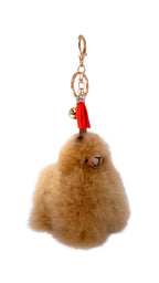 A fluffy light beige alpaca keychain charm with a small red tassel, gold keyring, and a tiny gold bell, shown on a plain white background.