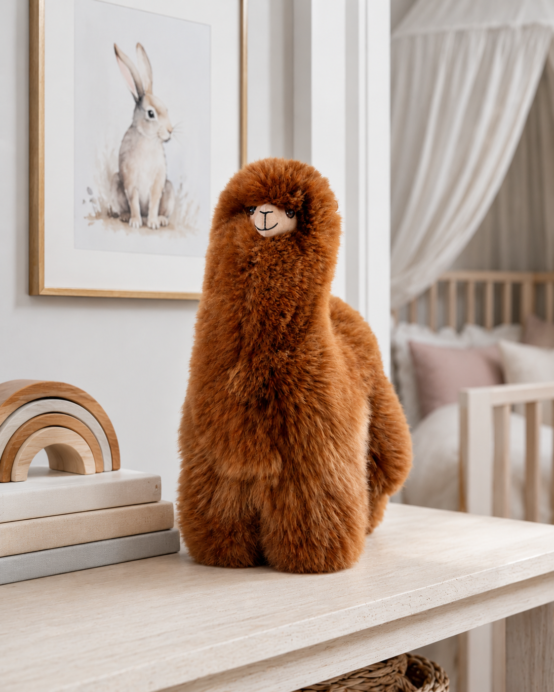 A fluffy brown alpaca plush toy sits on a light wood console table in a softly styled nursery. A framed watercolor rabbit print hangs on the wall behind it, with a crib and white canopy visible in the background.